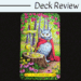 A tarot card from the Tarot of the Owls deck, The Magician shows an owl in a red cape among vibrant forest flowers, with a chalice, pentacle, wand, and sword on a tree stump. Deck Review appears in the background.