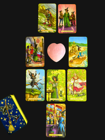 A tarot card spread with nine colorful cards arranged around a pink heart-shaped stone symbolizes lightening the load, with a tarot deck box featuring star patterns at the bottom left, all set against a black background.