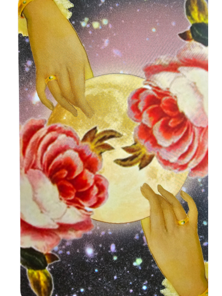 Two hands with gold rings hold a glowing full moon, surrounded by large red and pink flowers, against a starry night sky—a mystical scene inspired by The Rosebud Tarot.
