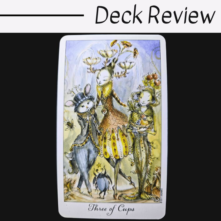 A whimsical tarot card from the Joie de Vivre Tarot labeled Three of Cups features three fanciful, animal-like characters in colorful attire, standing together against a soft, pastel background. Text above reads Deck Review on a white and black backdrop.