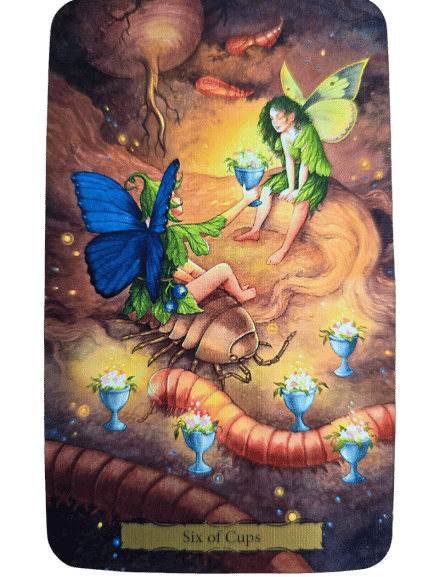 Two fairy-like creatures with colorful wings sit on a tree stump, smiling and offering cups filled with flowers. Six cups glow around them amid earthworms and lights, capturing the magical spirit of Tarot of the Witch's Garden. Text reads Six of Cups.