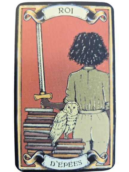 A tarot card from The Sacred Sisterhood Tarot, labeled Roi d’Épées, shows a curly-haired figure with their back turned, holding a sword. An owl perches on stacked books beside them against a reddish-orange background.